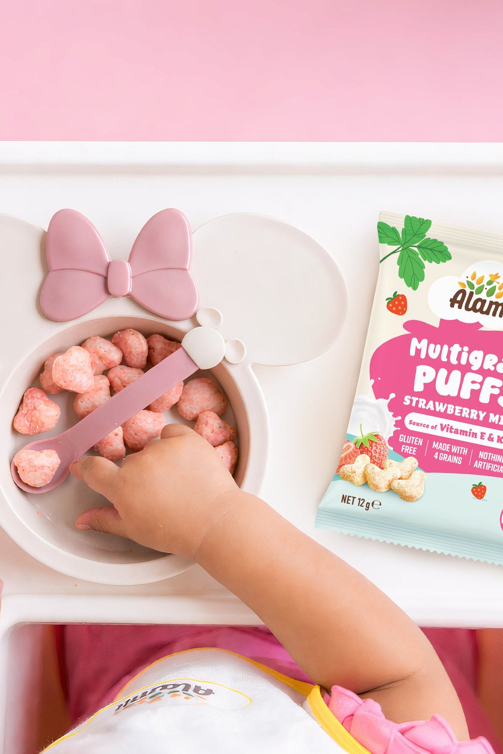 Child eating Alami Multigrain Puffs Strawberry Milk with a pink bowl and spoon on a white tray.